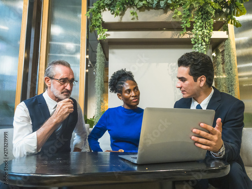 young male professional presents business project on laptop while his senior executive and colleague listen attentively inside modern corporate office meeting room.