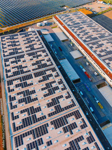 Solar panels on warehouse rooftops in industrial area near farmland. Workers and vehicles are seen at a large warehouse area with solar panels.