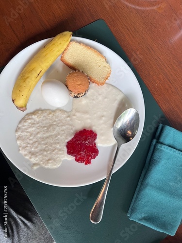 Canvastavla A white plate with a balanced breakfast including a banana, a piece of bread, a hard-boiled egg, and a creamy grain dish like porridge or rice pudding topped with a red fruit compote