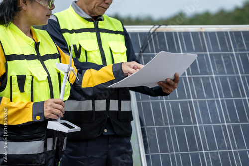 Two solar energy technicians in high visibility work wear inspect documents beside photovoltaic panels outdoors, checking a renewable power installation.