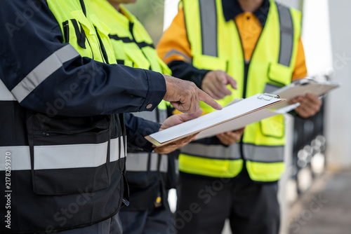 Construction workers in high visibility safety vests review documents on a clipboard during an on-site inspection and project meeting.