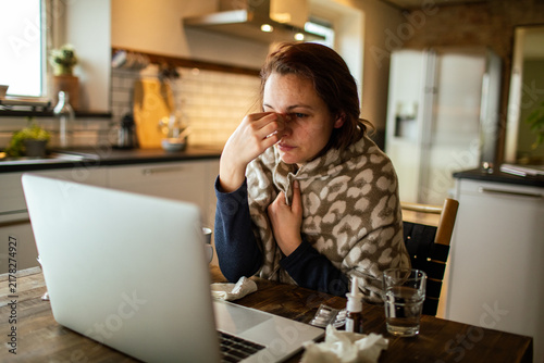 Ill woman wrapped in blanket working on laptop in home kitchen