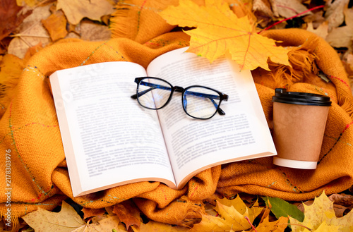 Cozy autumn still life with an open book, eyeglasses, coffee cup, warm blanket and colorful fallen leaves. Peaceful seasonal reading atmosphere. Selective focus.