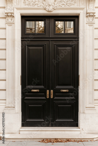 An ornate black double door with brass hardware stands within a cream stone facade, framed by classical architectural detailing.
