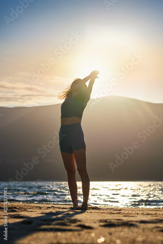 Backlit by the low sun, a woman practices an Urdhva Hastasana yoga variation, Upward Salute, with arms angled overhead beside sparkling beach water