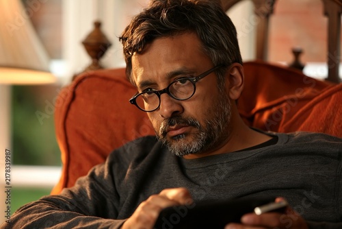 Thoughtful middle-aged man with glasses uses a tablet while relaxing in an armchair at home