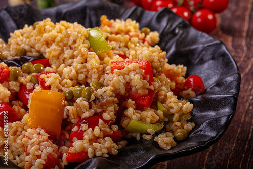 Fotografie Bulgur pilaf with mixed vegetables in black bowl on wooden table