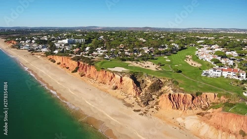 Scenic aerial footage showing the beautiful red cliffs of praia de garrao in vale do lobo, portugal, with a luxury golf course overlooking the turquoise atlantic ocean and sandy beach