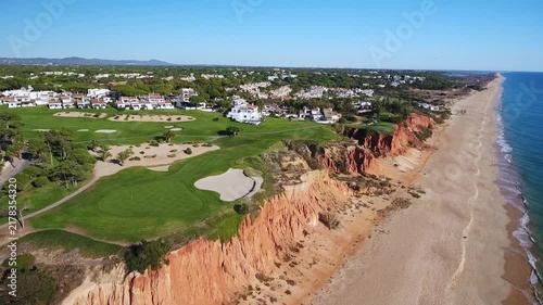 Beautiful aerial footage showing the stunning coastline of algarve, portugal, featuring a lush green golf course perched atop dramatic red cliffs next to a long sandy beach and the atlantic ocean