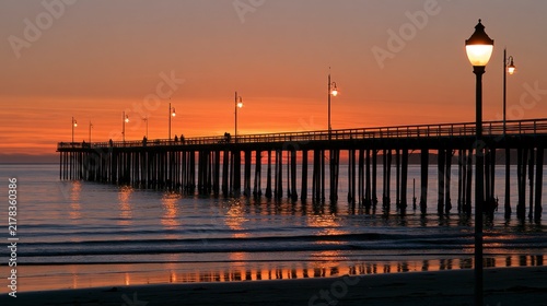 Scenic Wooden Pier Stretching into the Ocean during a Vibrant Orange Sunset with Glowing Street Lamps