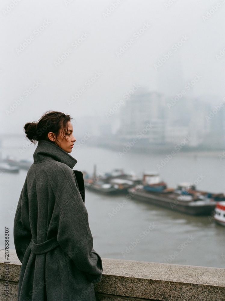 Fototapeta premium Young woman standing on a concrete wall overlooking a body of water. she is wearing a long grey coat and her hair is tied up in a bun.