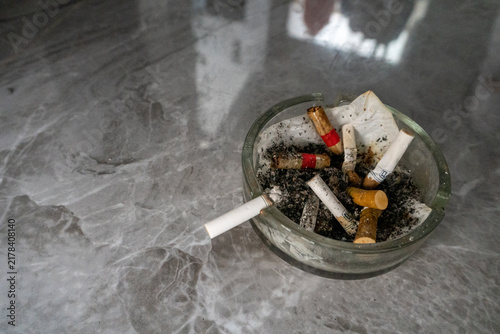 A clear glass ashtray containing used cigarette butts and gray ash sits on a gray surface with a specific pattern. An unlit or partially burnt cigarette protrudes over the edge of the ashtray.