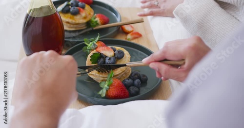 Foreground diner is placing fork and knife onto pancakes on bed tray, slicing bite to eat