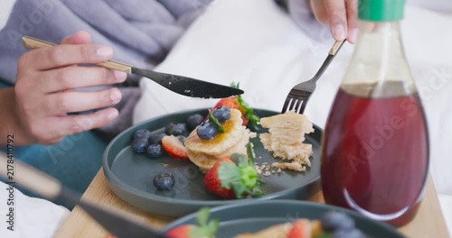 Knife is cutting pancakes on plate on bed tray as fork is lifting bite for eating