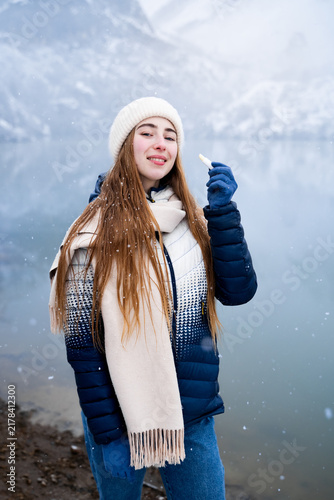 Foto Young woman in warm winter clothing applying moisturizing lip balm while looking at the camera against a snowy mountain lake, representing winter lip care and beauty skincare