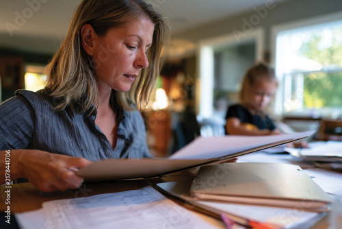 A dedicated mother is immersed in organizing documents at her home workspace while a child engages in nearby activities, highlighting family and productivity.