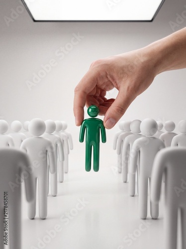 Human hand lifting green figurine from group of white standing figures on white surface with Hand picks up green toy figure crowd white plastic people standing rows recruitment