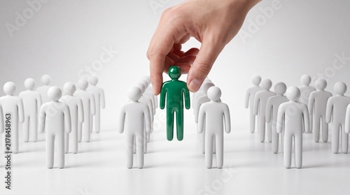 A hand picks out a green figure from a group of white people figurines with selects green toy people models figure crowd recruitment individual different selection isolation