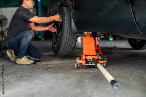 Selective focus to hydraulic floor jack with blurry professional mechanic installing a tire on a lifted car using an orange hydraulic floor jack and jack stand in a repair shop.