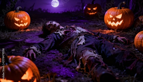 Undead zombie lying among glowing jack-o'-lanterns under full moon on Halloween night