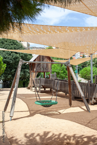 Basket swing and wooden playground equipment located in a public city park under shade sails surrounded by green trees. Outdoor recreational space designed for children, families and active leisure