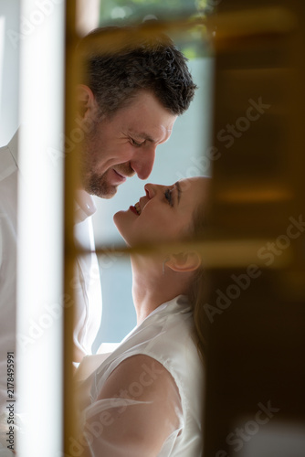 Romantic wedding portrait of a bride and groom sharing an intimate moment indoors, framed through a doorway. Soft natural light, genuine emotions, and timeless love.
