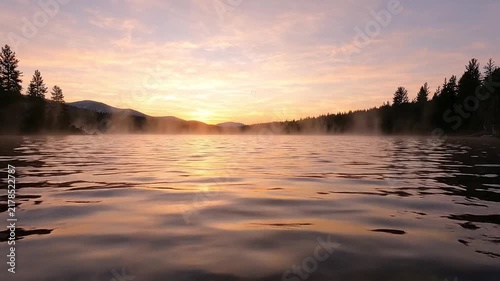 Misty lake surface with gentle ripples reflecting warm golden sunrise light over distant forest with while mist rises surface near distant shoreline trees mountains video calm lake