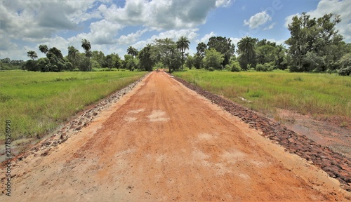 Vue de face d'une piste africaine en latérite rouge bordée de végétation. Route en terre rectiligne, avec un ciel nuageux à l'horizon. 