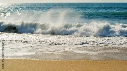 Ocean waves crashing onto a sandy beach shore during a bright sunny day at the coast with under bright sunlight breaking crashing video ocean onto wet environment with refreshing
