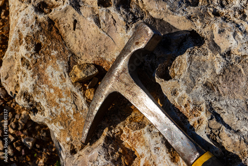 Steel geological hammer resting on a rough textured rock, professional rockhounding tool used for mineral exploration, excavation and stone collecting on a sunny outdoor field trip