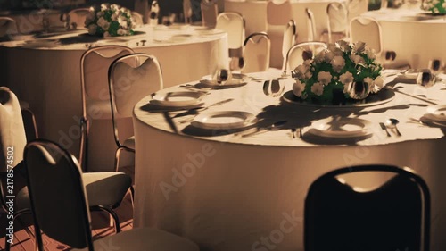 Tables are arranged with white tablecloths, dishes, and utensils ready for a formal dinner. Centerpieces of flowers sit on each table in the banquet hall. The room awaits guests.