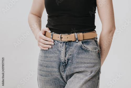 Close-up of a woman wearing a beige smooth leather belt with a classic metal buckle on acid wash jeans