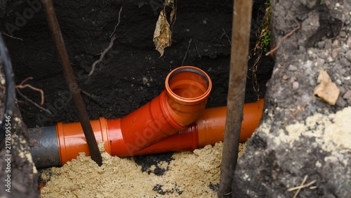 An orange plastic sewer pipe fitting installed in a deep dirt trench