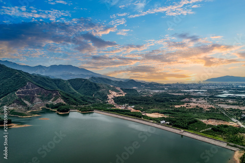 A lake bordered by green areas and mountains in Vietnam. Clouds cover the sky as the sun shines over the water and land
