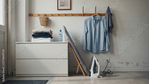 Stacked laundry with iron and ironing board standing near light blue shirt hanging on wooden wall rack for clothes care task.