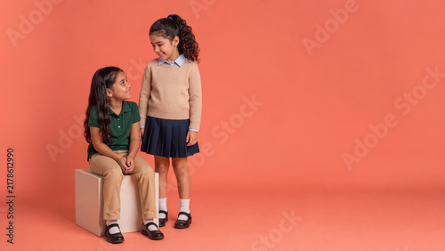 Two elementary schoolgirls model coordinated classroom outfits while smiling at each other, with one seated on a cube and the other standing in a coral studio.