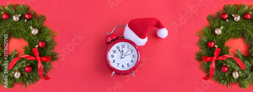 Alarm clock with Santa hat and Christmas wreath on a red background. Top view. Copy space.