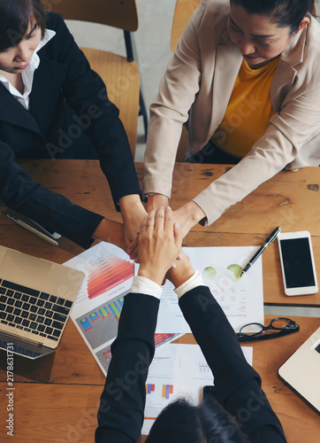 Group of businesswoman joining and hold in hands during business meeting at wooden table in indoor office, surrounded by laptops, tablets, documents and charts, showing teamwork, cooperation and unity