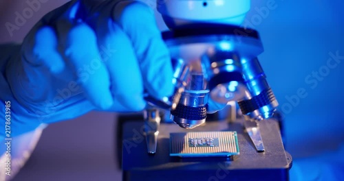 Tracking shot of gloved hand positioning microchip under microscope in laboratory close up with blue lighting and focusing on semiconductor component for inspection.