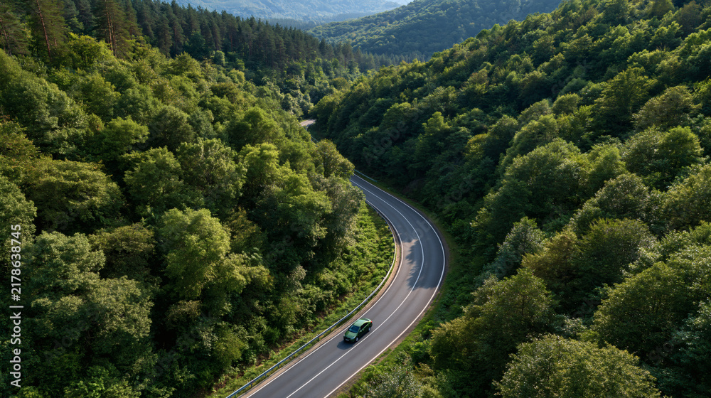 Fototapeta premium Winding Mountain Road: A solitary car navigates a scenic asphalt road that curves through a lush, verdant mountain landscape filled with dense evergreen trees.