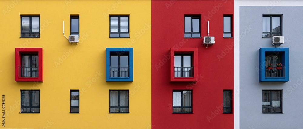 Obraz premium Symmetrical modern apartment building facade with solid blocks of bright yellow red and gray color accented with red and blue window frames showing bold contemporary geometric architectural design