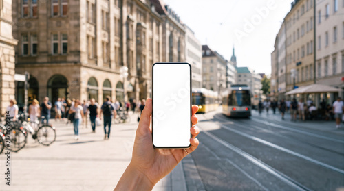 Hand holding a smartphone with a blank white screen mockup against a blurred sunny city street with pedestrians and a tram