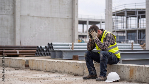 Worker sitting in despair holding his head at a heavy industrial construction site with concrete walls.