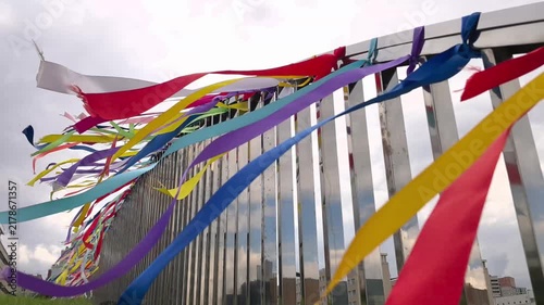 Colorful ribbons waving in wind on metal fence against cloudy sky. Rainbow streamers tied to railing fluttering in city. Festival decoration and wish making tradition concept.