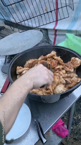Man cooking marinated chicken pieces in large cauldron outdoors. Hands putting grilled meat into big pot with tongs. Street food preparation, barbecue party catering concept.