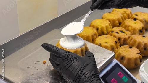 Baker in black gloves decorating small cake with white cream using spoon in commercial kitchen. Pastry confectioner preparing desserts with frosting, handmade bakery production concept