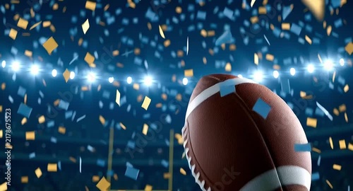Slow motion american football ball spinning and falling onto the green grass of a stadium at night. Confetti is raining down from above after a championship victory celebration
