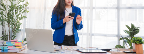Fotografie Businesswoman Working on Phone at Desk: A focused businesswoman in a blue blazer is engrossed in her smartphone while standing at a cluttered desk with a laptop, calculator, and documents