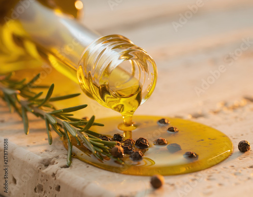Golden olive oil pouring from a clear bottle onto a light stone surface, with fresh rosemary and scattered whole black peppercorns
