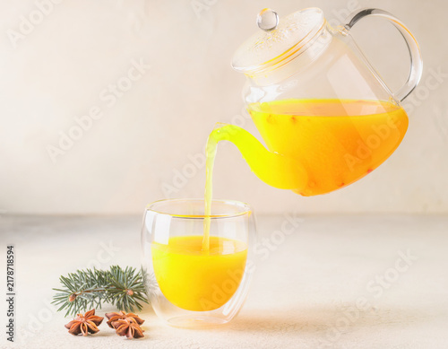 Vibrant sea buckthorn tea pours from a clear glass teapot into a modern double walled cup, with star anise and pine sprigs creating a cozy winter beverage